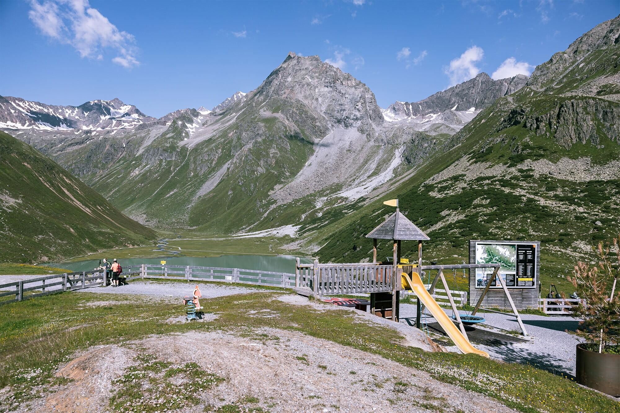 Ein Spielplatz in den Bergen mit einer Rutsche und einer Schaukelturm. Im Hintergrund sind grüne Hügel und hohe Berge zu sehen.