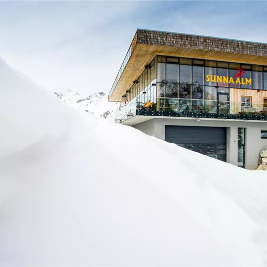 Ein modernes Gebäude in einer verschneiten Berglandschaft. Die Fassade ist mit großen Fenstern und einem Balkon ausgestattet.