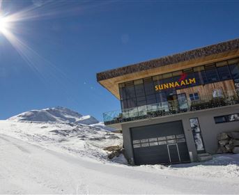Ein modernes Gebäude in den Bergen, umgeben von Schnee. Die Sonne scheint klar in den blauen Himmel.
