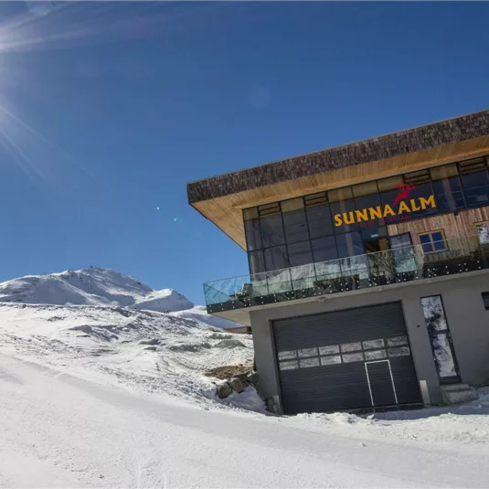 Ein modernes Gebäude in den Bergen, umgeben von Schnee. Die Sonne scheint klar in den blauen Himmel.