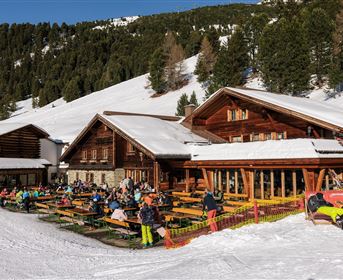 Eine alpine Hütte im Schnee mit vielen Gästen, die draußen an Holztischen sitzen. Die Umgebung ist von schönen Bergen und Bäumen umgeben.
