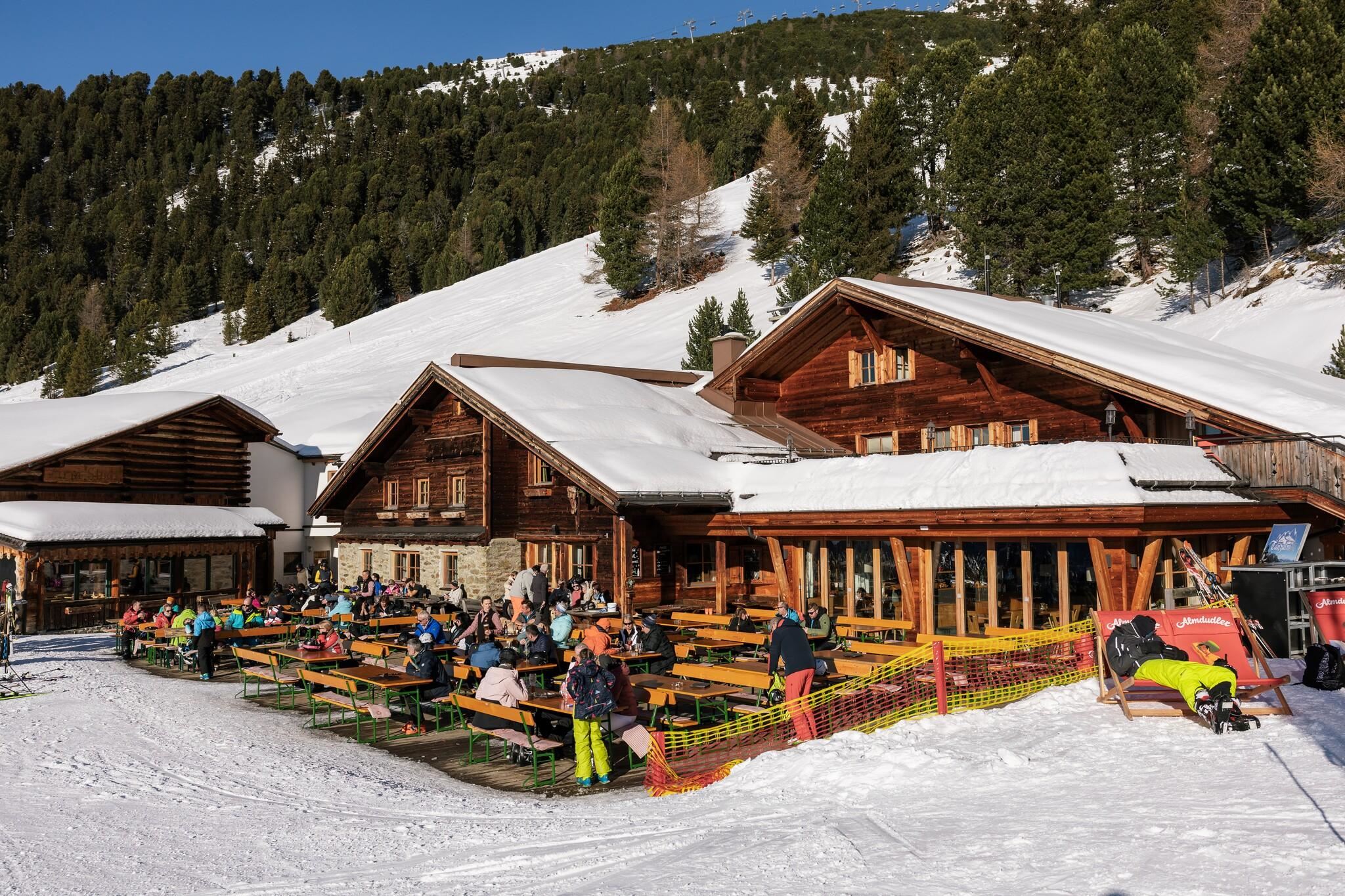 Eine alpine Hütte im Schnee mit vielen Gästen, die draußen an Holztischen sitzen. Die Umgebung ist von schönen Bergen und Bäumen umgeben.