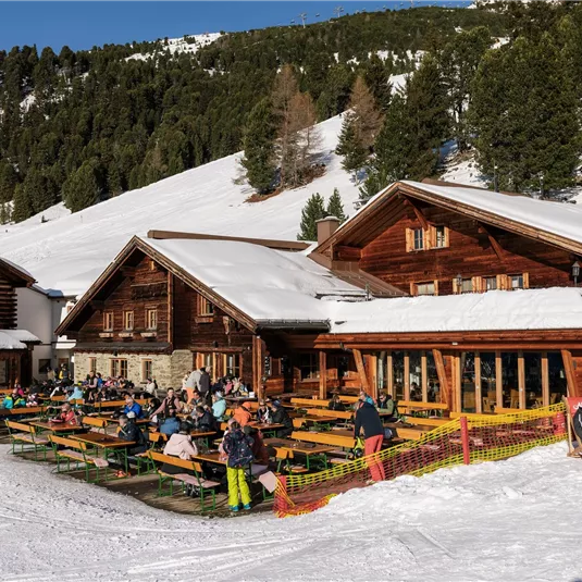 Eine alpine Hütte im Schnee mit vielen Gästen, die draußen an Holztischen sitzen. Die Umgebung ist von schönen Bergen und Bäumen umgeben.