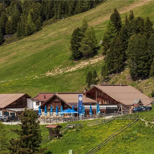 Ein alpenländliches Gasthaus mit Terrassen im Freien und Sonnenschirmen. Umgeben von grünen Wiesen und hohen Tannenbäumen.