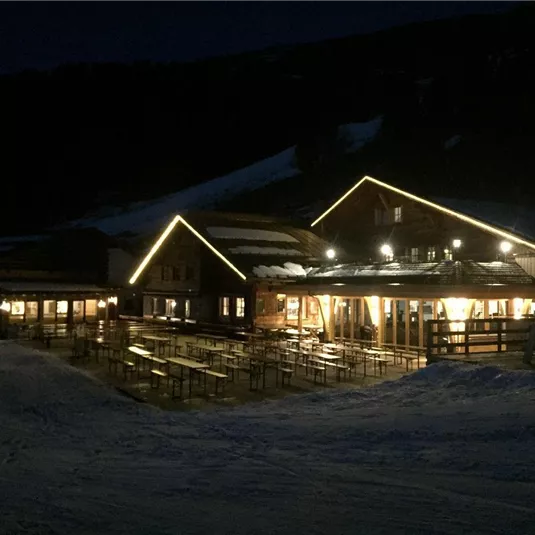 Ein beleuchtetes Restaurant in einer winterlichen Landschaft. Es gibt viele Tische im Freien und die Umgebung ist schneebedeckt.