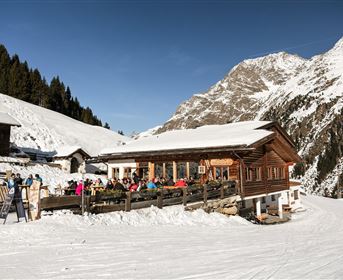 Eine gemütliche Hütte in den Bergen, umgeben von Schnee. Gäste genießen die Aussicht auf die winterliche Landschaft.