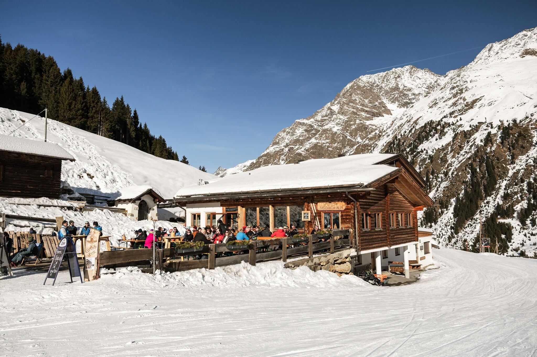 Eine gemütliche Hütte in den Bergen, umgeben von Schnee. Gäste genießen die Aussicht auf die winterliche Landschaft.