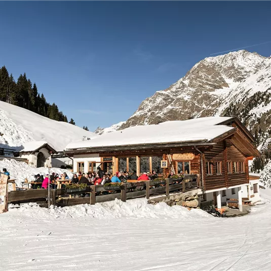 Eine gemütliche Hütte in den Bergen, umgeben von Schnee. Gäste genießen die Aussicht auf die winterliche Landschaft.