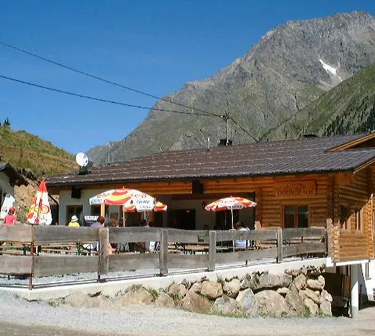Eine gemütliche Berghütte mit Terrasse und Sonnenschirmen. Im Hintergrund sind beeindruckende Berge zu sehen.