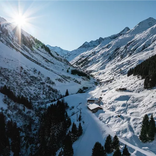 Eine schneebedeckte Berglandschaft mit einem klaren Himmel und strahlender Sonne. Im Tal sind Bäume und eine kleine Hütte zu sehen.