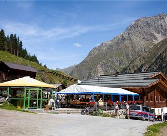 A mountain hut with several terraces and tables. In the background, high mountains and a blue sky can be seen.