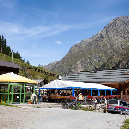 A mountain hut with several terraces and tables. In the background, high mountains and a blue sky can be seen.