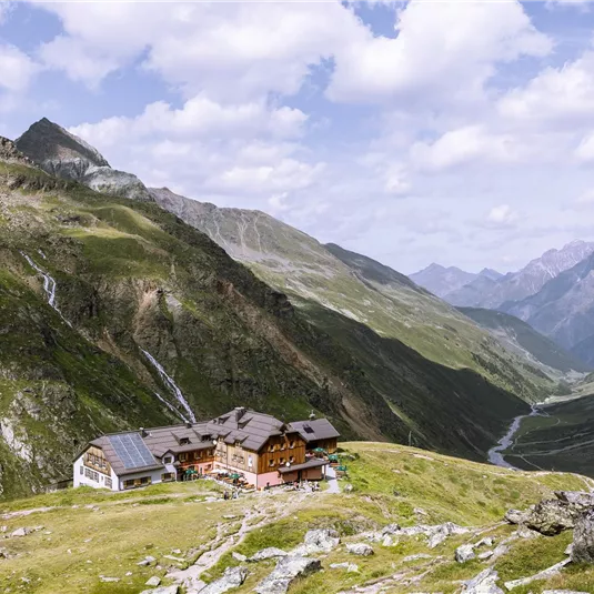 Eine malerische Berglandschaft mit sanften Hügeln und einem klaren Himmel. Im Vordergrund steht ein rustikales Haus, umgeben von grünen Wiesen und schroffen Felsen.