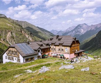 Ein gemütliches Berggasthaus umgeben von grünen Wiesen und hochgelegenen Bergen. Der Himmel ist teilweise bewölkt mit einigen sonnigen Stellen.