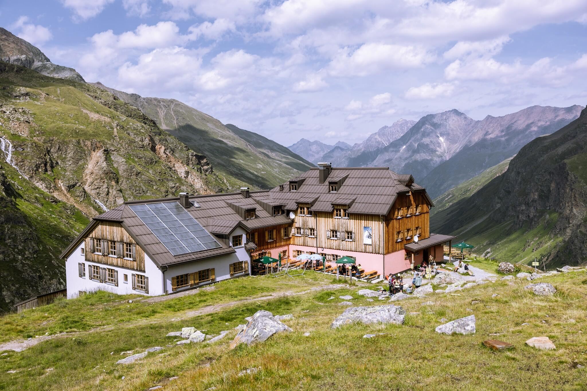 Ein gemütliches Berggasthaus umgeben von grünen Wiesen und hochgelegenen Bergen. Der Himmel ist teilweise bewölkt mit einigen sonnigen Stellen.