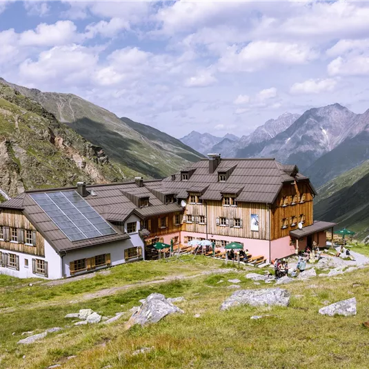 Ein gemütliches Berggasthaus umgeben von grünen Wiesen und hochgelegenen Bergen. Der Himmel ist teilweise bewölkt mit einigen sonnigen Stellen.