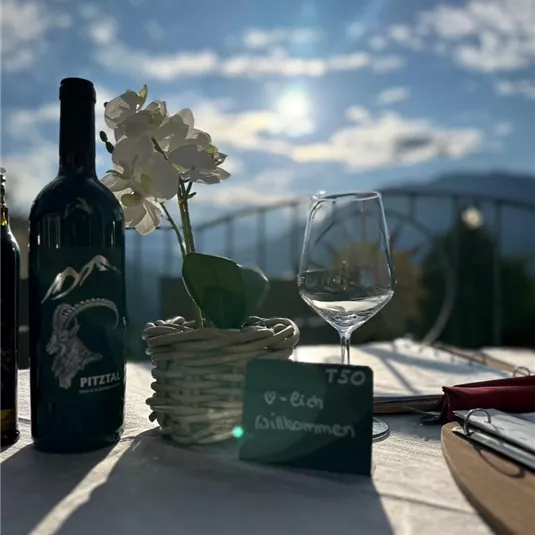 A set table with a bottle of wine, a glass, and a blooming plant. In the background, there are mountains and a clear sky.