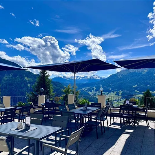 A beautiful terrace with tables and chairs, surrounded by mountains and a clear blue sky. Umbrellas provide shade and invite you to linger.