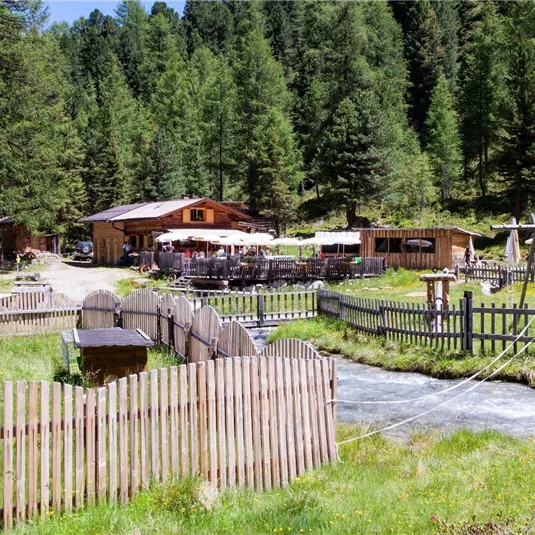 Ein idyllisches Restaurant inmitten von Wäldern, umgeben von saftigem Grün. Im Vordergrund fließt ein klarer Bach, und es gibt einen Spielplatz für Kinder.