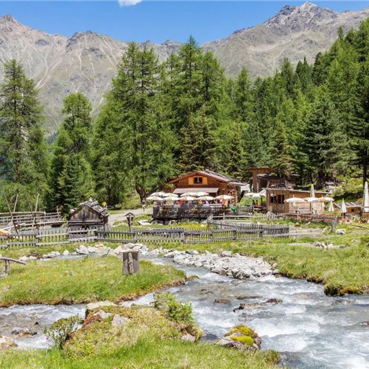 Eine malerische Alpenlandschaft mit einem klaren Fluss und üppigen, grünen Wäldern. Im Hintergrund sind Berge und gemütliche Hütten zu sehen.