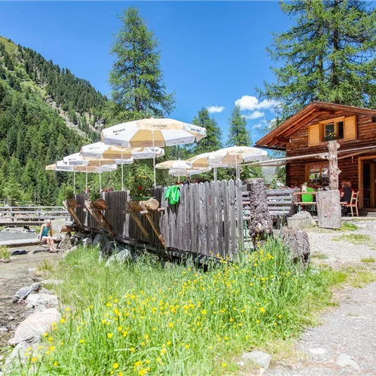 Eine gemütliche Almhütte inmitten von grünen Wiesen und Bäumen. Sonnenschirme bieten Schatten, während ein kleiner Bach vorbeifließt.
