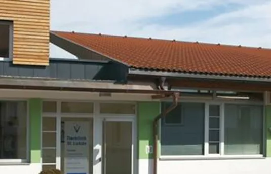 Ein modernes Gebäude mit einem Flachdach und großen Fenstern. Die Fassade ist teilweise weiß gestrichen und hat grüne Akzente.