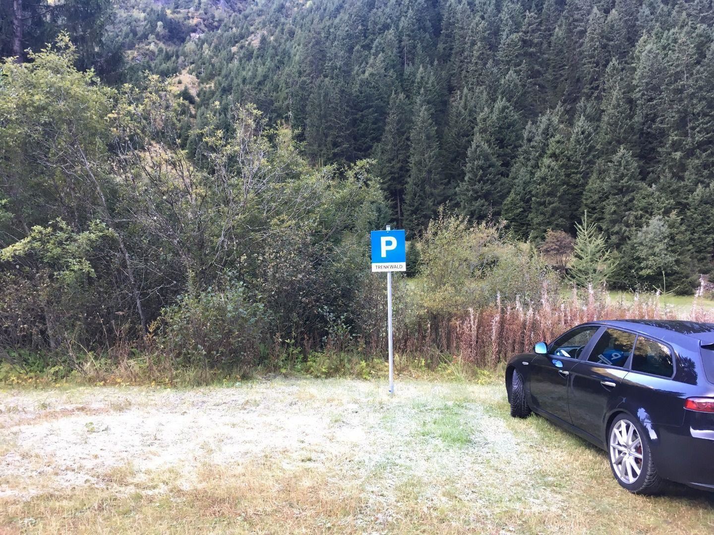 Ein geparktes Auto neben einem Parkplatzschild in einer ländlichen Umgebung. Im Hintergrund sind Bäume und Hügel zu sehen.