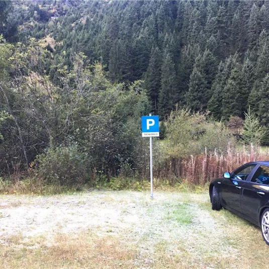 Ein geparktes Auto neben einem Parkplatzschild in einer ländlichen Umgebung. Im Hintergrund sind Bäume und Hügel zu sehen.