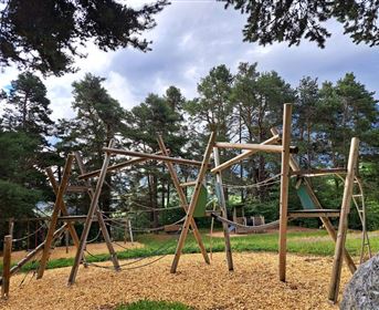 Eine Spielplatzstruktur aus Holz mit Seilbrücken und Kletterelementen. Im Hintergrund sind hohe Bäume und ein bewölkter Himmel zu sehen.