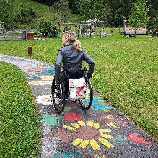 A girl in a wheelchair is riding along a colorful path painted with flower motifs. In the background, green meadows and trees can be seen.