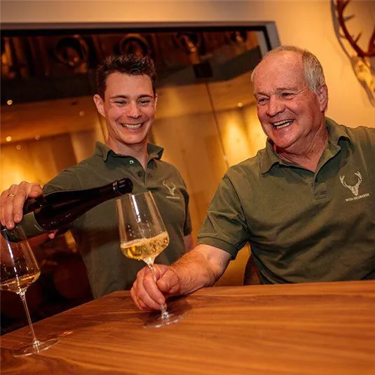 Zwei Männer stehen an einem Tisch und gießen Sekt in Gläser. Beide haben ein Lächeln im Gesicht und tragen grüne Polo-Shirts.
