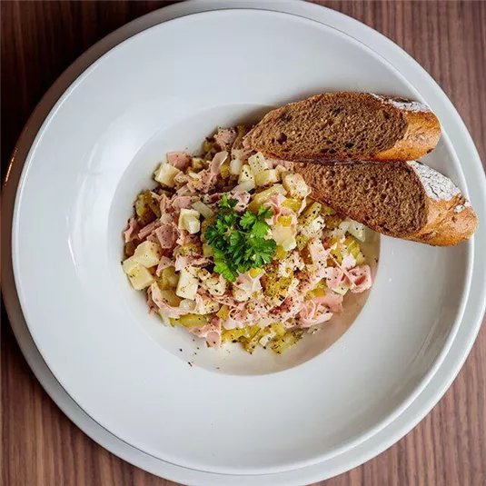 Ein Teller mit einem gemischten Salat aus Kartoffeln, Schinken und einer cremigen Soße. Dazu gibt es ein Stück Brot.