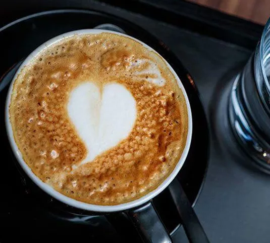 Eine Tasse Kaffee mit Herz-Muster aus Milchschaum auf einem schwarzen Unterteller. Neben der Tasse steht ein Glas Wasser.