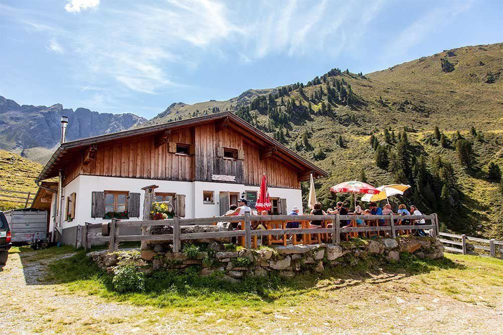 Eine gemütliche Berghütte mit einer Terrasse voller Gäste. Im Hintergrund sind grüne Hügel und blauer Himmel zu sehen.