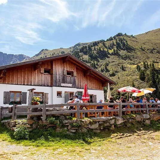 Eine gemütliche Berghütte mit einer Terrasse voller Gäste. Im Hintergrund sind grüne Hügel und blauer Himmel zu sehen.