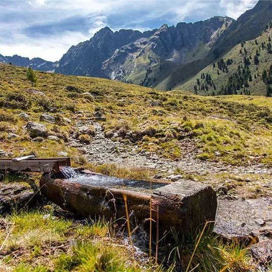 Ein ruhiger Gebirgsbach fließt über ein Holzgestell in einer grünen Landschaft. Im Hintergrund sind majestätische Berge zu sehen.