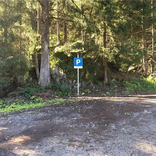 Ein Parkplatzschild steht am Rand eines Waldgebiets. Die Umgebung ist grün und von Bäumen umgeben.