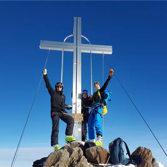 Drei Bergsteiger stehen auf einem Gipfel neben einem großen Kreuz. Sie feiern ihren Erfolg unter einem klaren blauen Himmel.