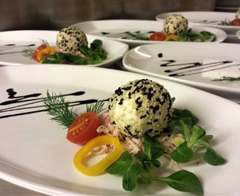 Ein elegant angerichteter Teller mit einem Reisball, der mit schwarzem Sesam bestreut ist. Daneben liegen frische Kräuter, Tomaten und bunte Paprika.