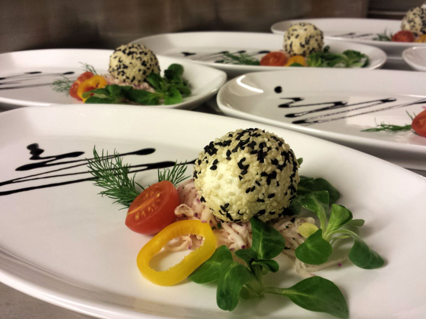 Ein elegant angerichteter Teller mit einem Reisball, der mit schwarzem Sesam bestreut ist. Daneben liegen frische Kräuter, Tomaten und bunte Paprika.