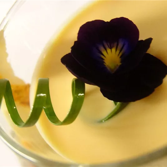 Eine cremige Dessertschicht in einem Glas, garniert mit einer essbaren Blume und einer spiralförmigen Dekoration. Das Bild vermittelt einen eleganten und ansprechenden Eindruck.