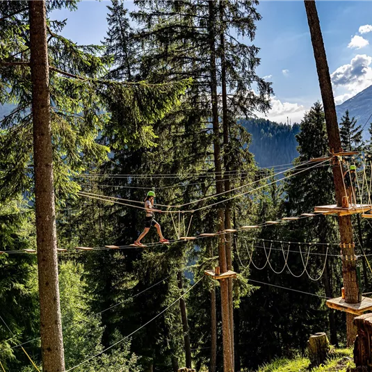 Ein Hochseilgarten inmitten von hohen Bäumen. Eine Person balanciert auf einer Slackline über dem Boden.