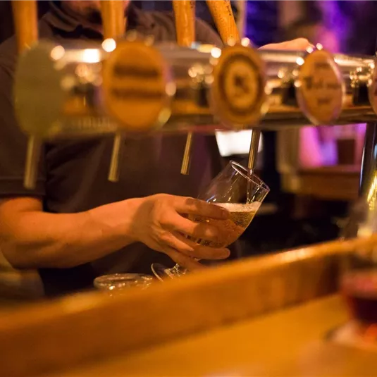 Ein Barkeeper zapft ein Bier in ein Glas. Die warme, gedämpfte Beleuchtung schafft eine gemütliche Atmosphäre.