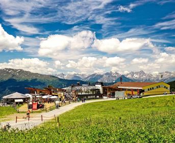 A picturesque mountain view with green meadows and a modern building. In the background, impressive mountains and a blue sky with a few clouds can be seen.