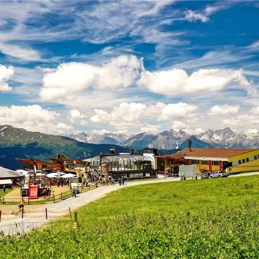 A picturesque mountain view with green meadows and a modern building. In the background, impressive mountains and a blue sky with a few clouds can be seen.