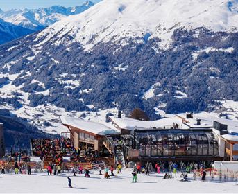 Eine Skiregion mit starkem Schnee und einem großen Berghaus. Viele Skifahrer genießen die winterliche Landschaft und die Berge im Hintergrund.