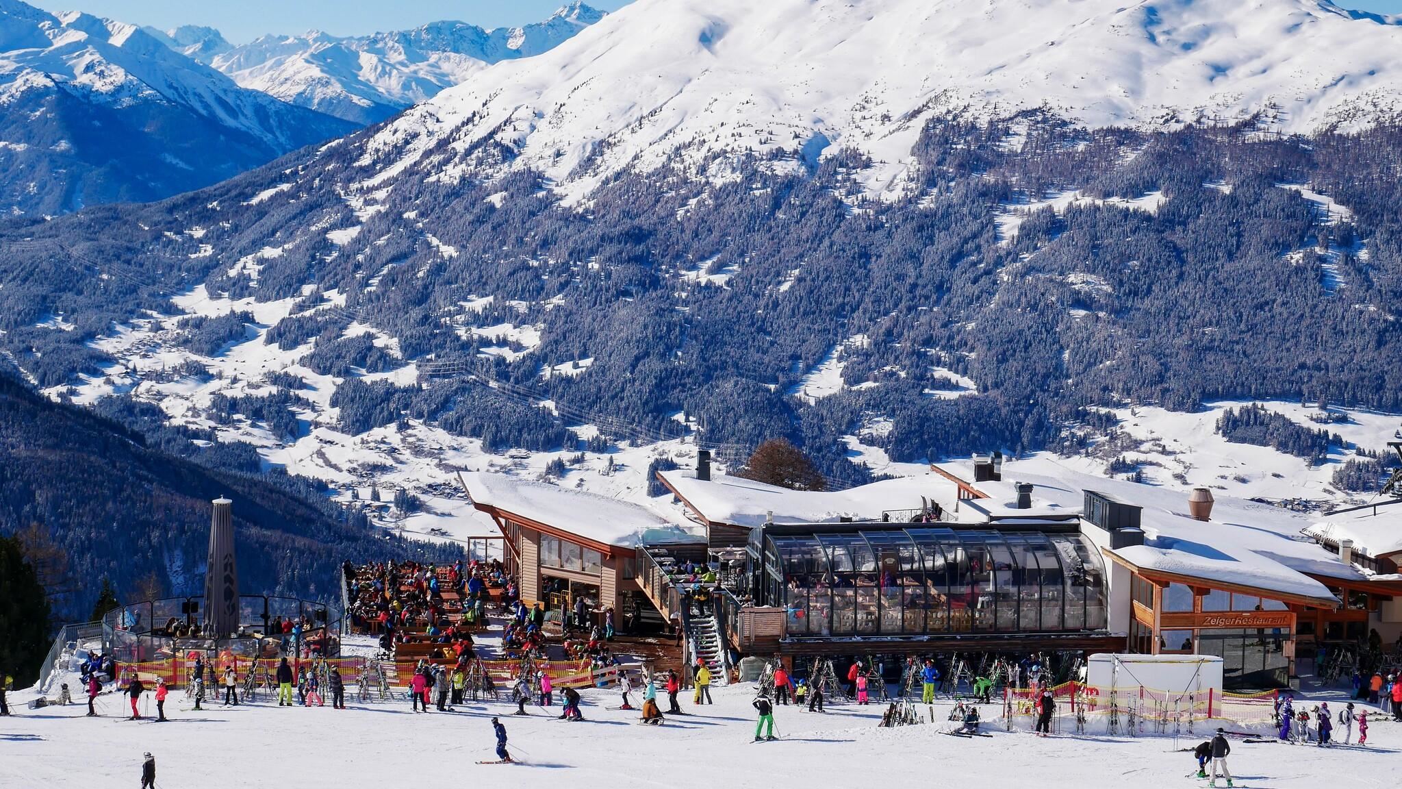 Eine Skiregion mit starkem Schnee und einem großen Berghaus. Viele Skifahrer genießen die winterliche Landschaft und die Berge im Hintergrund.