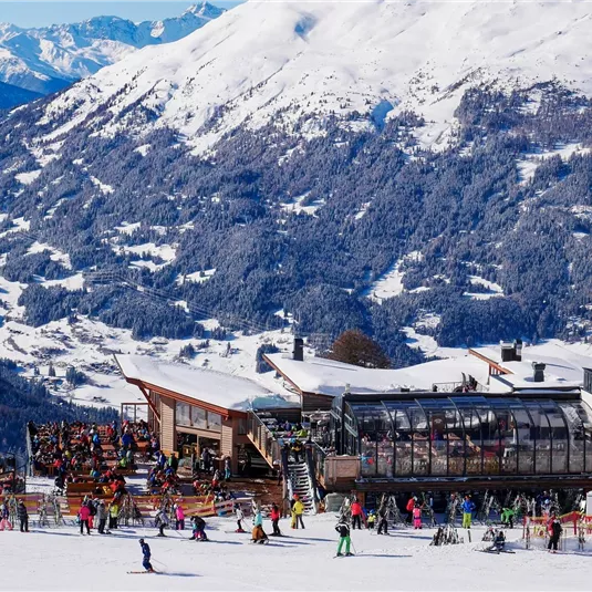 Eine Skiregion mit starkem Schnee und einem großen Berghaus. Viele Skifahrer genießen die winterliche Landschaft und die Berge im Hintergrund.