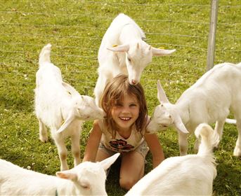 A smiling girl sits on the grass, surrounded by several small goats. The scene conveys joy and innocence in a rural setting.