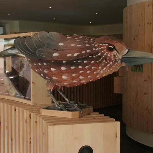 Eine große Vogelskulptur aus Holz steht in einem modernen Raum. Die Skulptur hat eine detailreiche Bemalung und ist auf einem Holzsockel platziert.